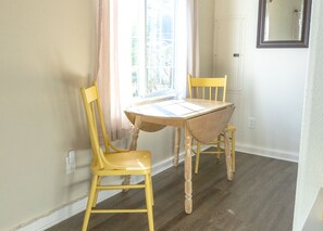 A room with a wooden table and two yellow chairs, a window with curtains, and a door with a mirror on the wall.
