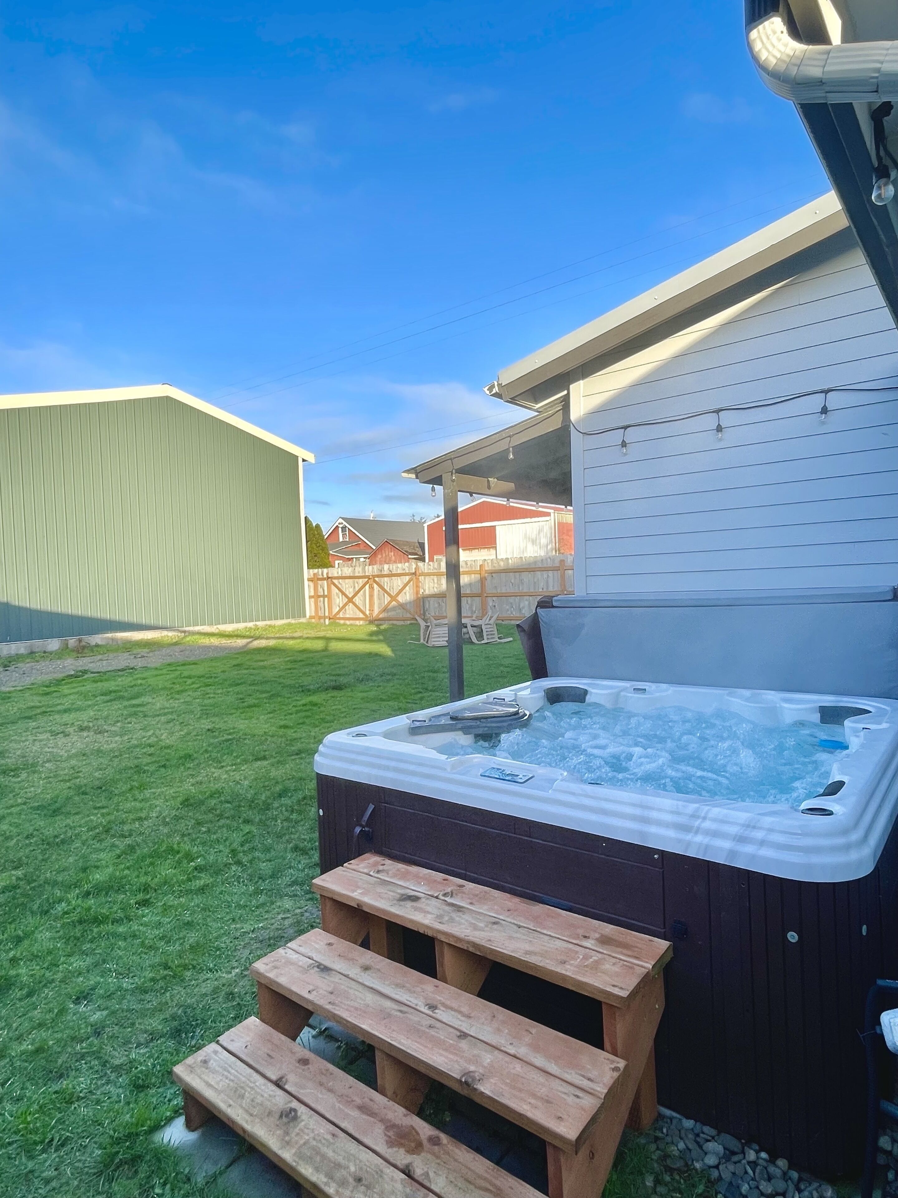A hot tub with a wooden bench in front, set in a backyard with a grassy lawn and a wooden fence.
