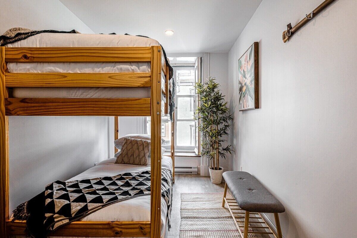 A bunk bed with a geometric-patterned blanket, a potted plant, and a bench in a narrow room with a window and a painting on the wall.