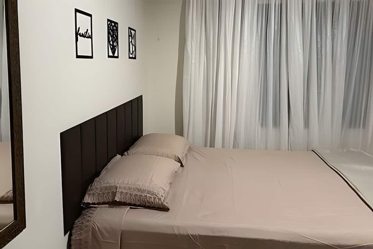 A bed with a dark headboard, a beige bedspread, and a window with sheer curtains.