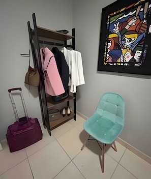 Una habitación con una silla color turquesa, un armario de madera con ropa colgada, una maleta morada y una obra de arte enmarcada en la pared.