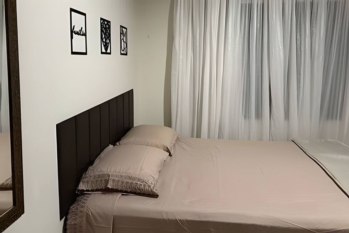 Un lit avec une tête de lit sombre, une couvre-lit beige et une fenêtre ornée de rideaux légers.