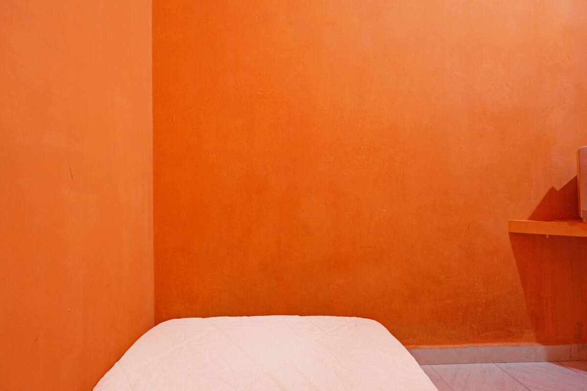 A room with an orange wall, a bed with a white mattress, and a small wooden nightstand.