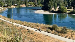 Une rivière, avec un cycliste sur une piste cyclable asphaltée, entourée d’arbres et avec des montagnes en arrière-plan.