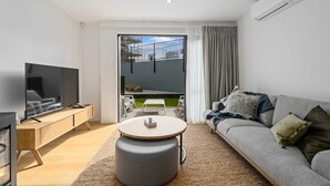 A modern living room with a flat-screen TV, a wooden sideboard, a grey sofa, and a round ottoman.