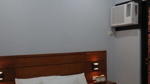 A hotel room with a wooden headboard, a bed with white linens, a bedside table with a lamp, a wall-mounted air conditioner, and a trash bin. - Cerca Hotel (Boracay Island)