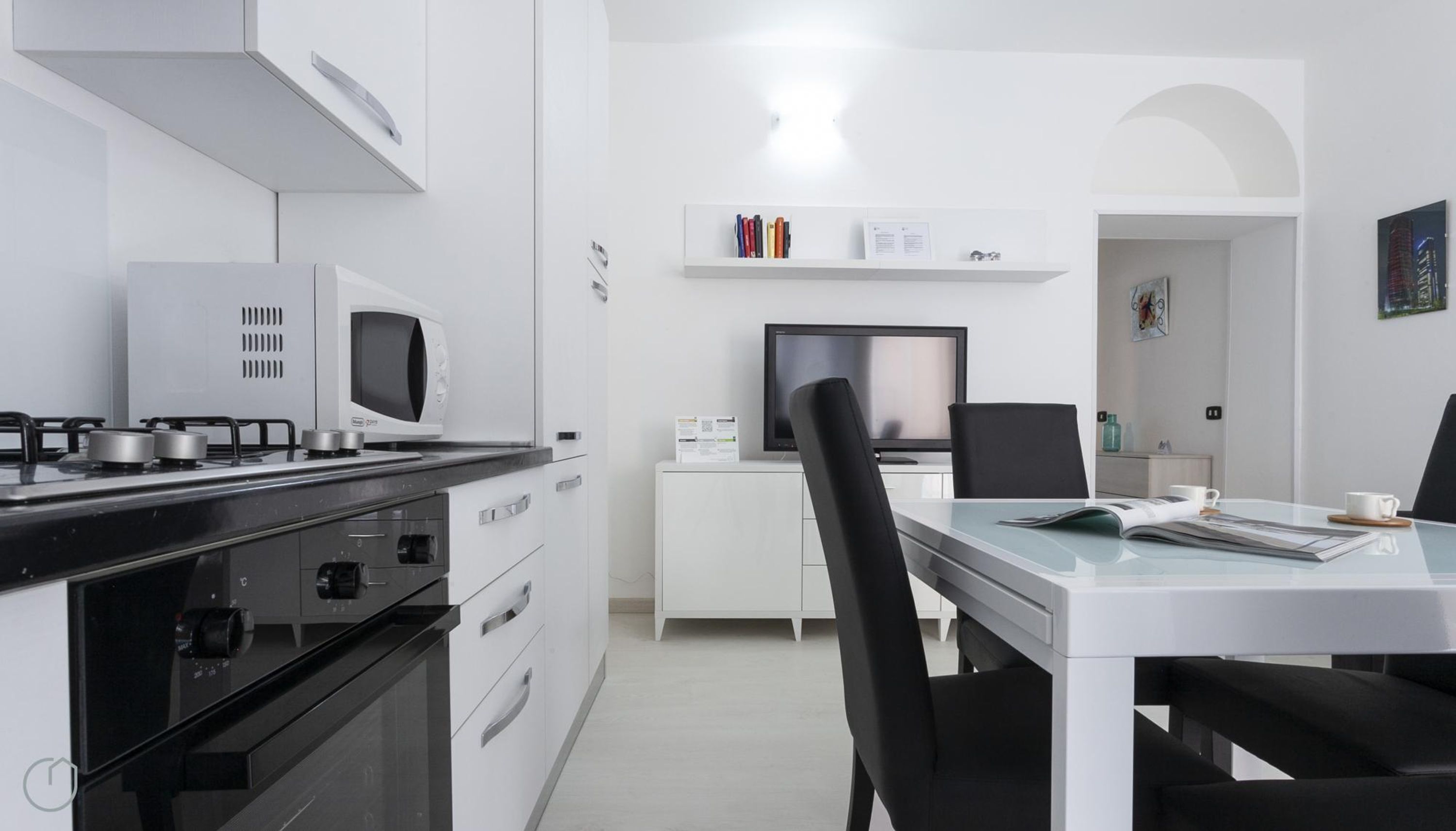 A modern kitchen with a gas stove, microwave, and oven, a dining area with a glass table and black chairs, and a small TV on a white cabinet.