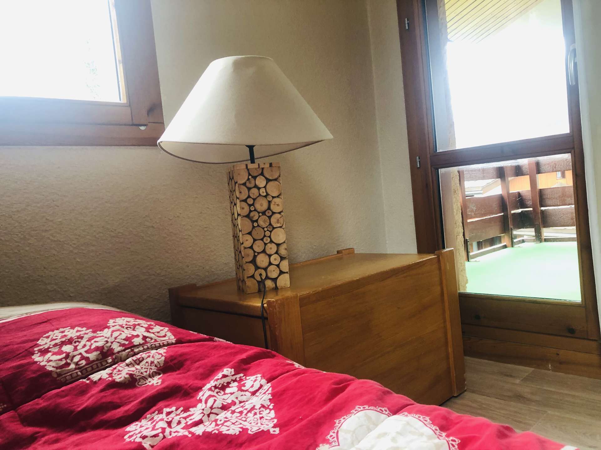 Ein Schlafzimmer mit Bett, Nachttisch mit Lampe und Fenster mit Blick auf eine Holzterrasse.