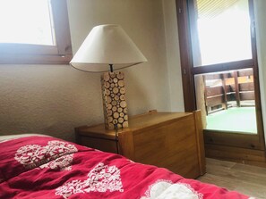 A bedroom with a bed, a bedside table with a lamp, and a window with a view of a wooden deck. - Teppes 46 - Cosy duplex ski in, ski out (LES AVANCHERS VALMOREL)