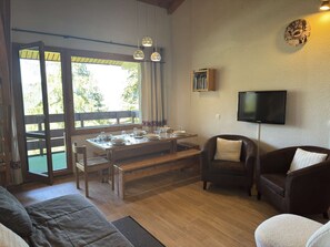 A cozy living room with a wooden dining table set for four, a sofa, a leather armchair, and a television mounted on the wall. - Teppes 46 - Cosy duplex ski in, ski out (LES AVANCHERS VALMOREL)