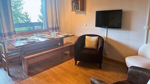 Un comedor con una mesa de madera preparada para dos comensales, un sillón de cuero con un cojín, un sofá y un televisor de pantalla plana montado en la pared.
