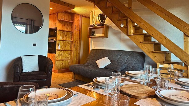 Una sala de estar acogedora con una escalera de madera, un sofá, una mesa de comedor preparada para cuatro personas y una chimenea.