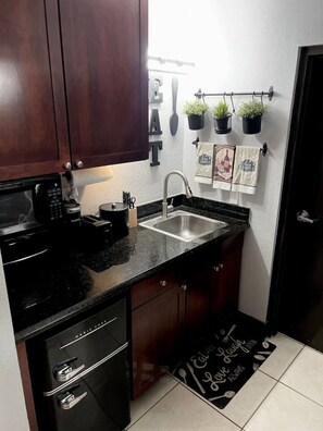 A compact kitchen with dark wooden cabinets, a black countertop, a stainless steel sink, and a microwave. There are potted plants on the wall and a kitchen mat with a message on the floor.