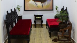 Ein Zimmer mit einem großen Portrait an der Wand, einem Deckenventilator, einer hölzernen Bank mit rotem Kissen und einem kleinen Tisch mit einer Pflanze.