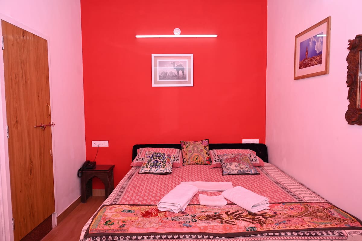 A room with a red wall, a bed with patterned bedding, a wooden door, and a small bedside table with a lamp.