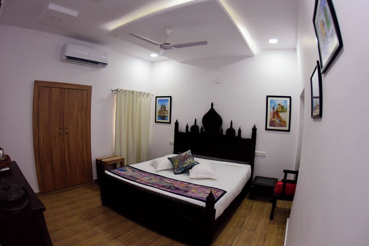 A bedroom with a bed, wooden wardrobe, ceiling fan, air conditioning unit, and framed pictures on the wall.