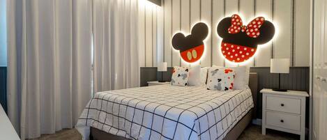 A modern bedroom with a large bed, Mickey Mouse head-shaped wall lights, striped wallpaper, and a small bedside table.