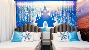 Quarto de hotel com duas camas, cada uma com almofada decorada com flocos de neve, uma mesinha de cabeceira e uma tapeçaria retratando um castelo.