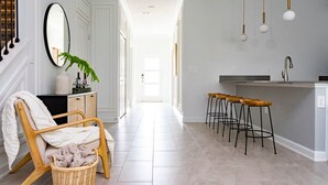 Corredor que dá para uma cozinha com espaço de bar, uma cadeira de vime com um cobertor e um espelho pendurado.