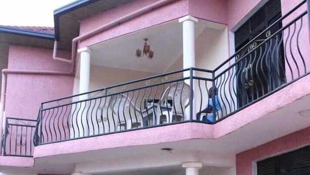 Un edificio de dos pisos con fachada rosa, un balcón con barandilla negra y un patio con baldosas.