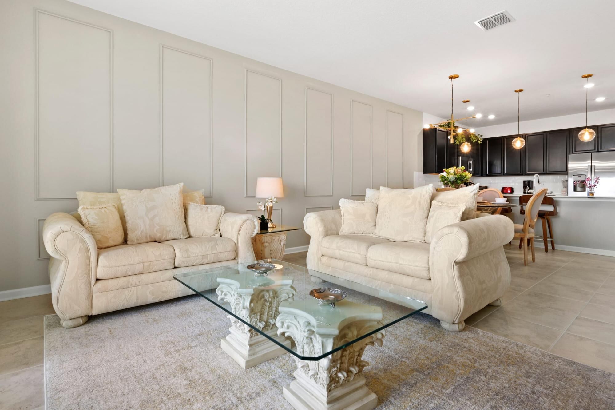 Ein Wohnzimmer mit zwei hellbeigen Sofas, einem Glastisch und einer Küche im Hintergrund.