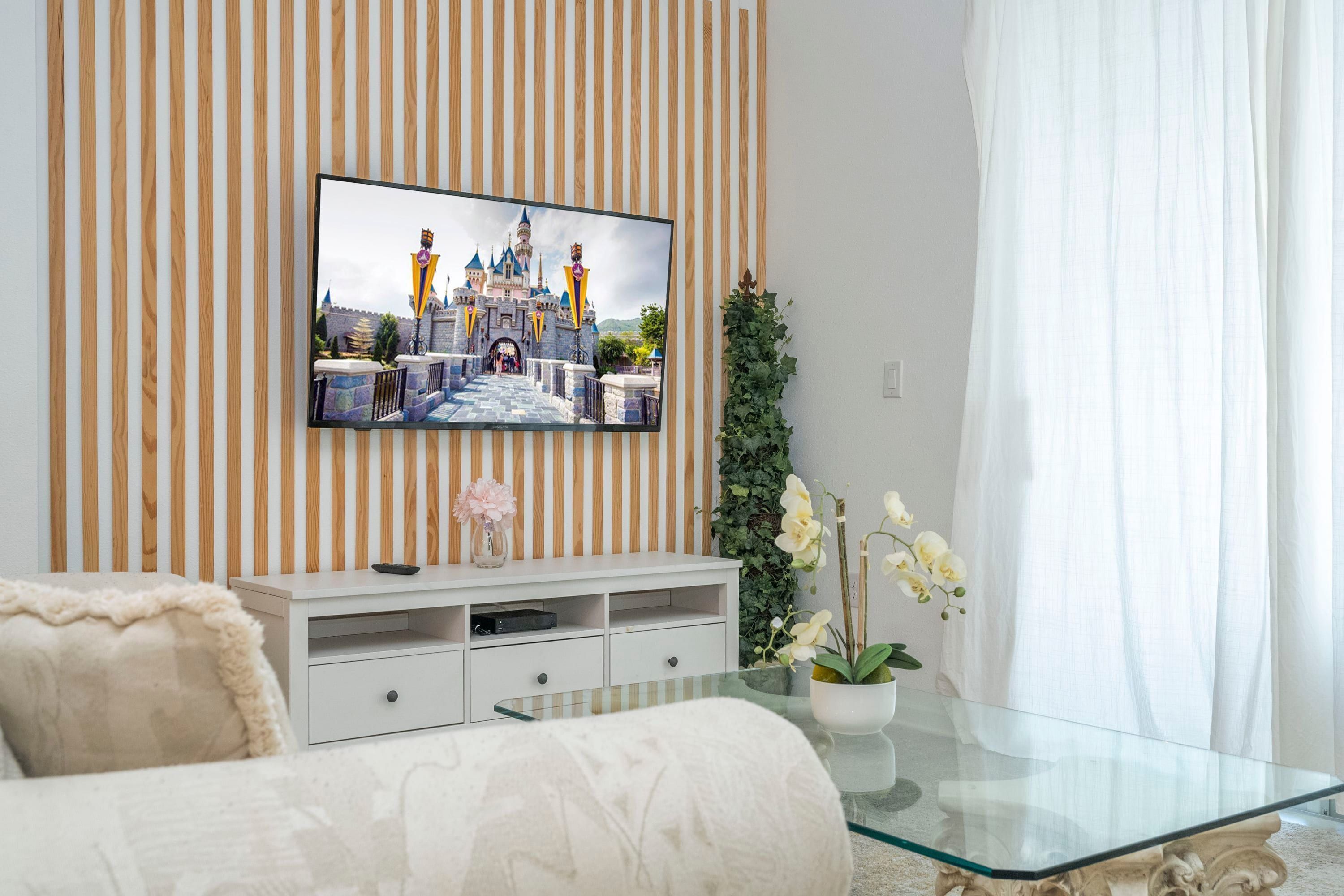 A modern living room with a flat-screen TV mounted on a wooden wall, a glass coffee table, a white sofa, and a small white cabinet.