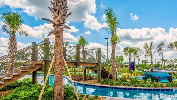 Un espace piscine agrémenté de palmiers, d’un pont en bois et d’un ciel d’un bleu limpide.