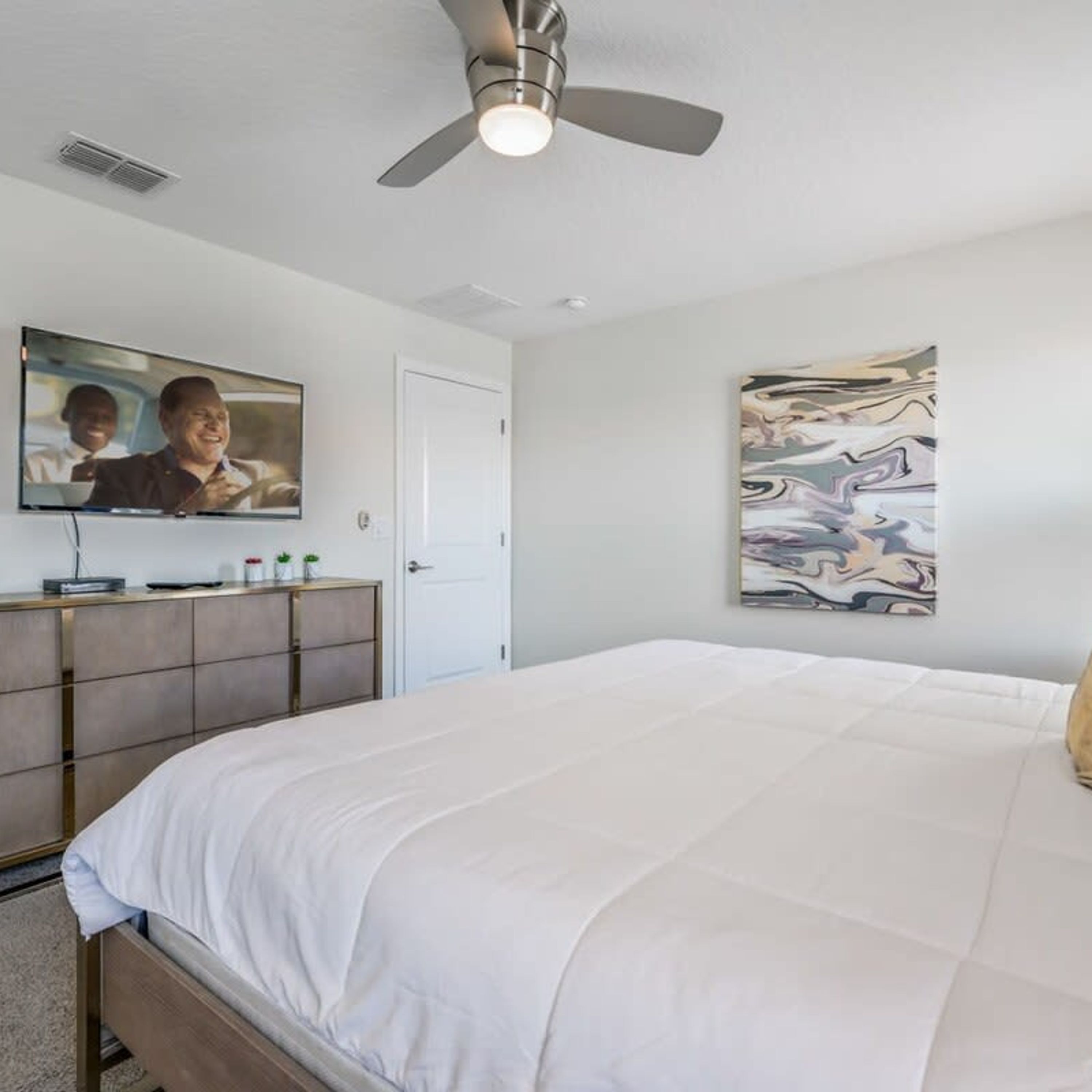 Un dormitorio con cama, ventilador de techo, televisor montado en la pared, una puerta y un cuadro.