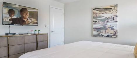 Un dormitorio con cama, ventilador de techo, televisor montado en la pared, una puerta y un cuadro.