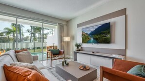 Un salon moderne doté d’un grand téléviseur à écran plat, d’un canapé, d’une table basse et offrant une vue sur une piscine et la verdure environnante.