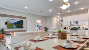 Un comedor moderno con una mesa larga dispuesta para una comida, un sofá y una cocina al fondo.