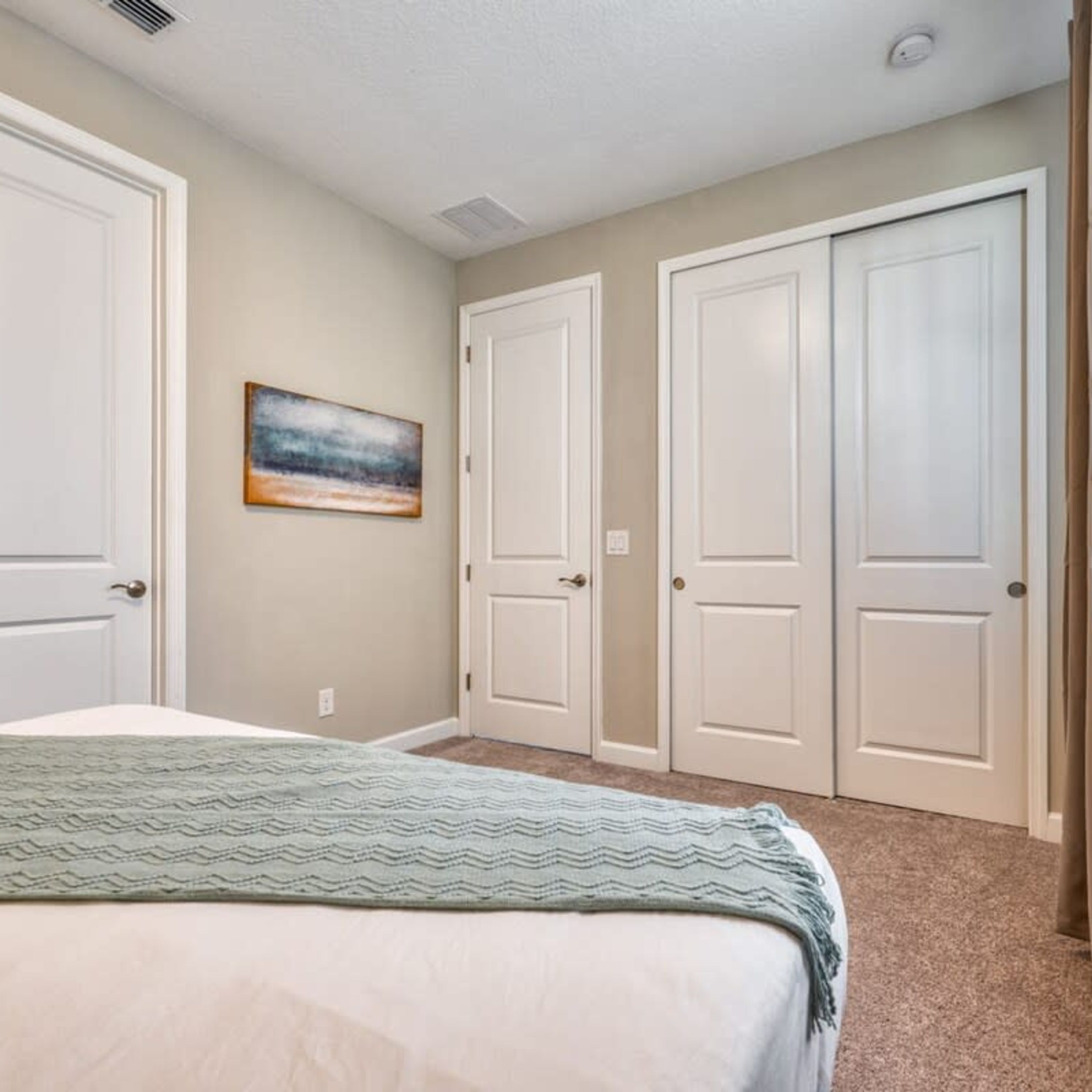 A bedroom with a bed, two white doors, a painting on the wall, and a carpeted floor.