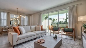 Uma sala de estar moderna com sofá, mesa de centro e cadeiras. Há uma área de jantar com mesa e cadeiras. Janelas amplas oferecem vista para palmeiras e um cercado.