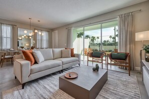 A modern living room with a sofa, coffee table, and chairs. There is a dining area with a table and chairs. Large windows provide a view of palm trees and a fence. - Luxury 5 Bedroom Home 10 Minutes From Disney (Kissimmee)