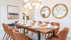 A dining area with a long wooden table set for six, surrounded by brown leather chairs. There is a chandelier with multiple light bulbs above the table, and two round mirrors on the wall.