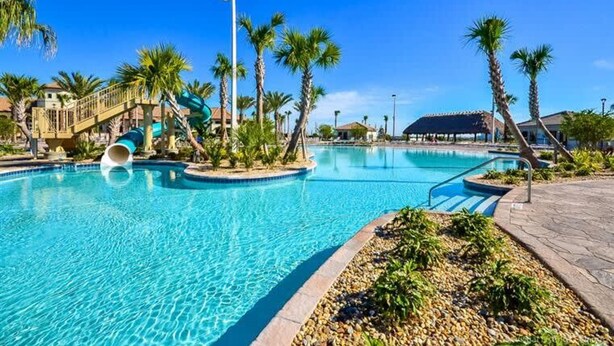 A swimming pool with a water slide, palm trees, and a covered area in the background.