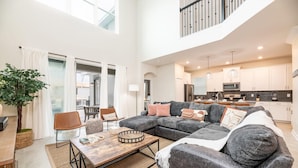 A spacious living area with a large sectional sofa, a wooden coffee table, and a dining table. There is a kitchen in the background with modern appliances and cabinets.