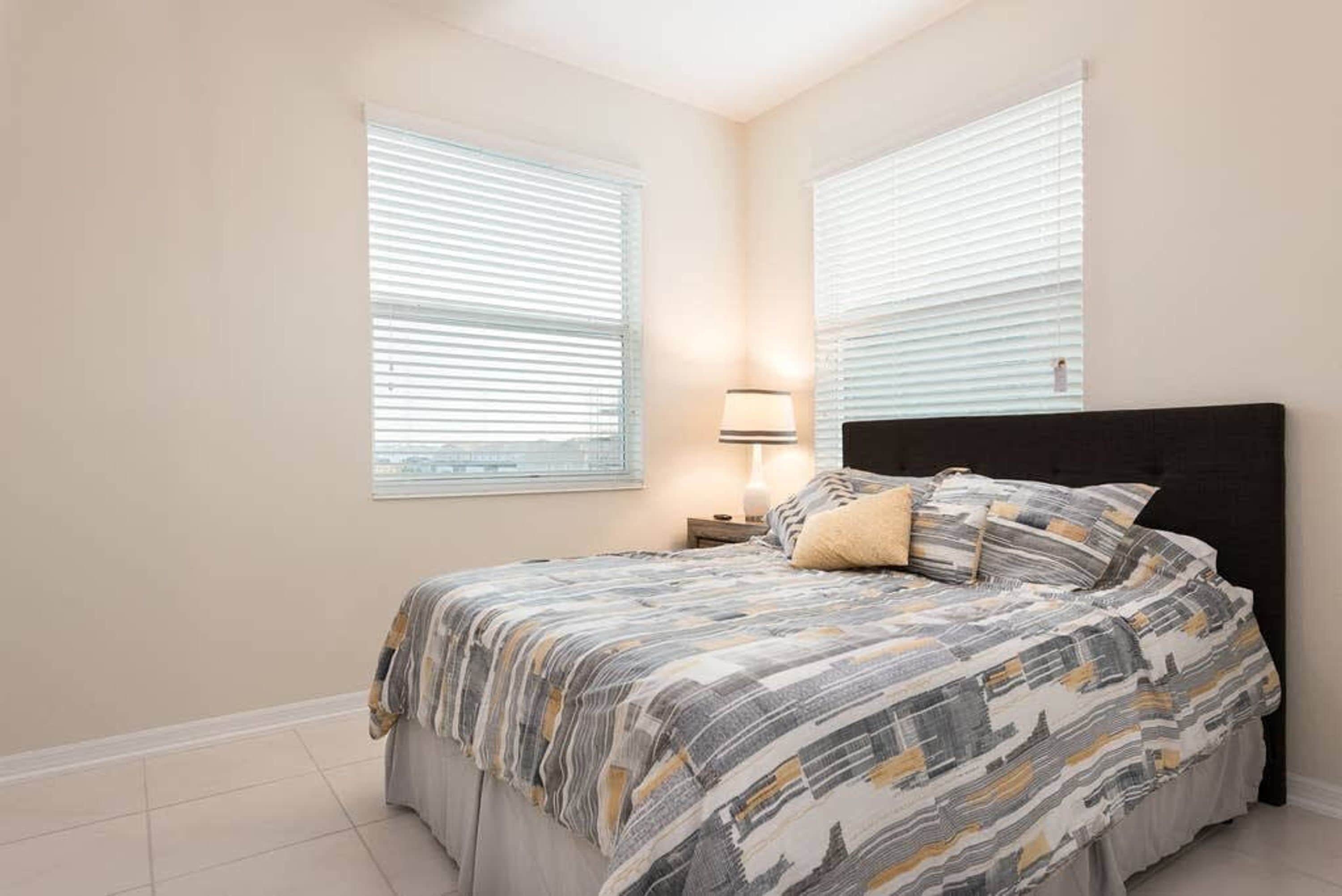 A bedroom with a bed, two bedside tables, a lamp, and two windows with blinds.