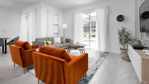 Ett modernt vardagsrum med en grå soffa, orange fåtöljer, ett soffbord och ett stort fönster med gardiner.