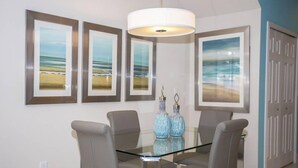 A dining area with a glass table and four chairs, framed ocean landscape paintings on the wall, and a door in the background. - Charming Cozy Haven in Four Corners (Clermont)