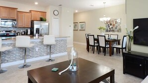 A modern living room with a coffee table, a dining area with a chandelier, and a kitchen with wooden cabinets and appliances. - Charming 4 BDR Townhome at Storey Lake Resort (Kissimmee)