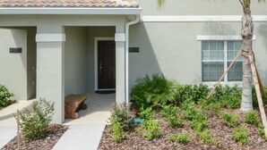 A two-story building with a tiled roof, a covered porch, and a small garden area with various plants and a palm tree. - Charming 4 BDR Townhome at Storey Lake Resort (Kissimmee)