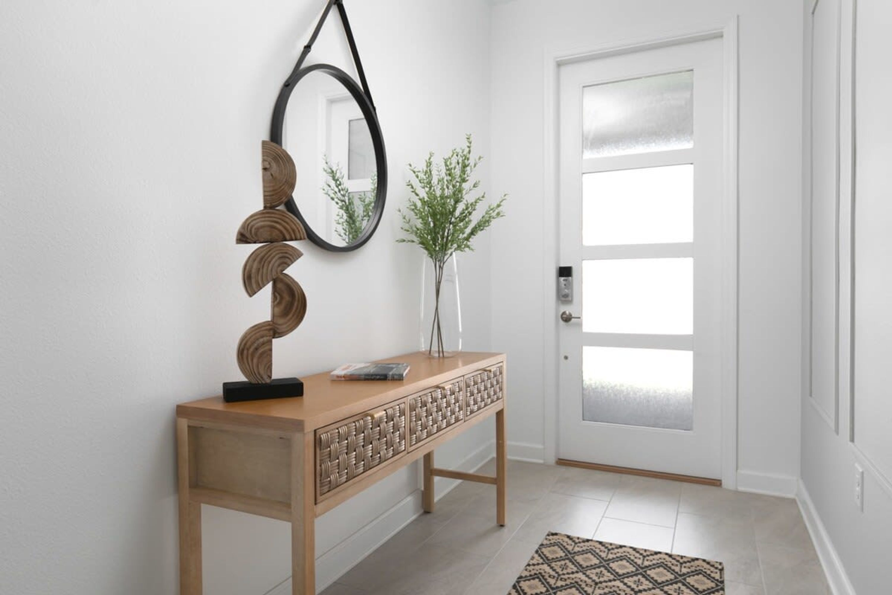 A wooden console table with a mirror and decorative items, a door with frosted glass panels, and a patterned rug on a tiled floor.