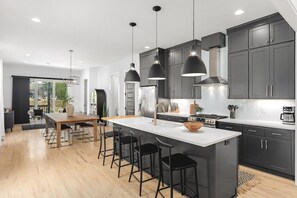 A modern kitchen with dark cabinets, a central island with stools, pendant lighting, and a dining area with a wooden table and chairs.