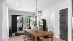 A modern dining area with a wooden table and metal chairs, a hanging light fixture, and a large window offering a view of the outdoors. - Huge Rooftop Patio at this Stunning Spacious House (Nashville)