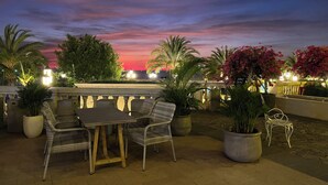 Un espace extérieur aménagé avec une table et des chaises, entouré de plantes en pot et de palmiers, offrant une vue sur le coucher du soleil.