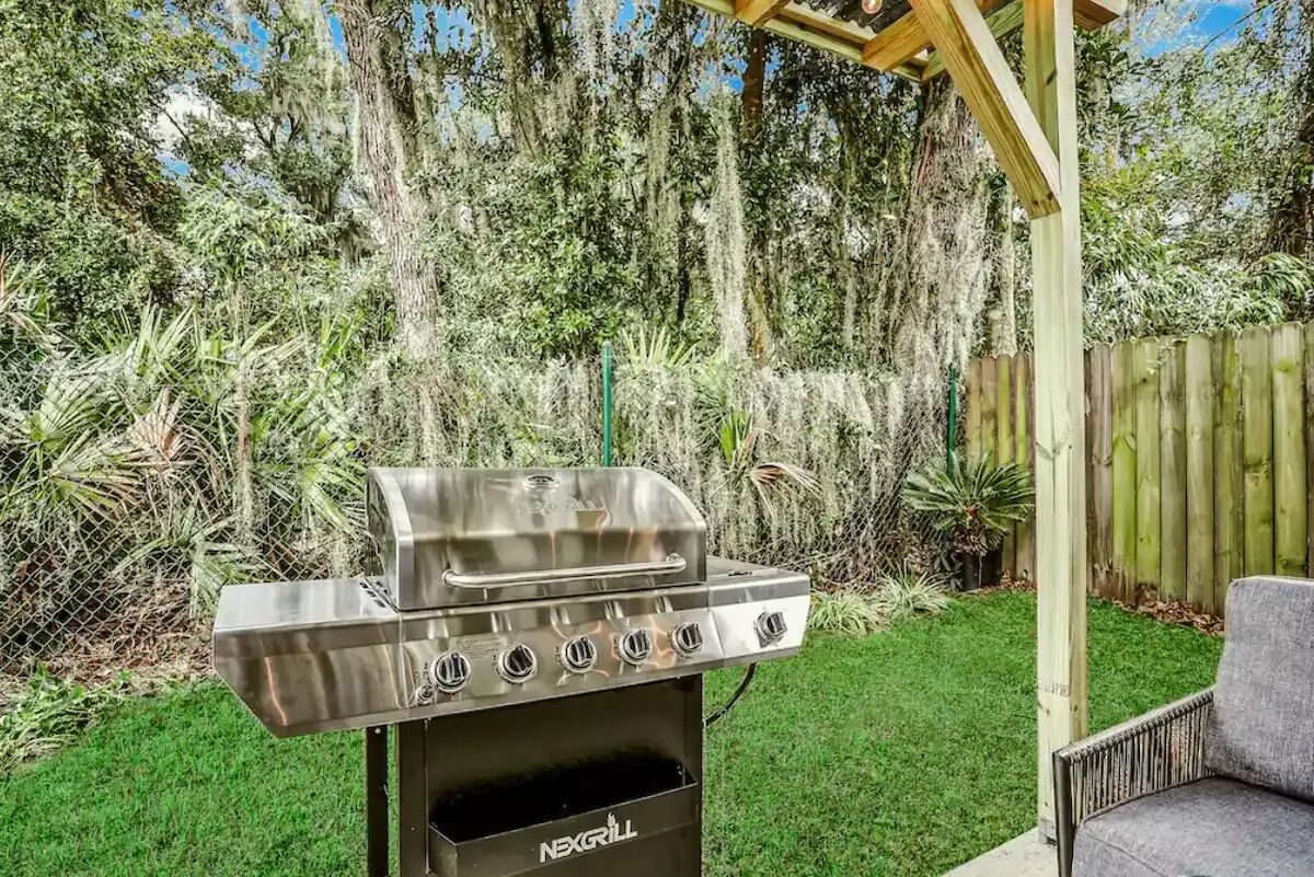 Una parrilla de acero inoxidable con cuatro quemadores y un compartimento de cocción negro, ubicada en un patio trasero con cerco de madera y diversas plantas.