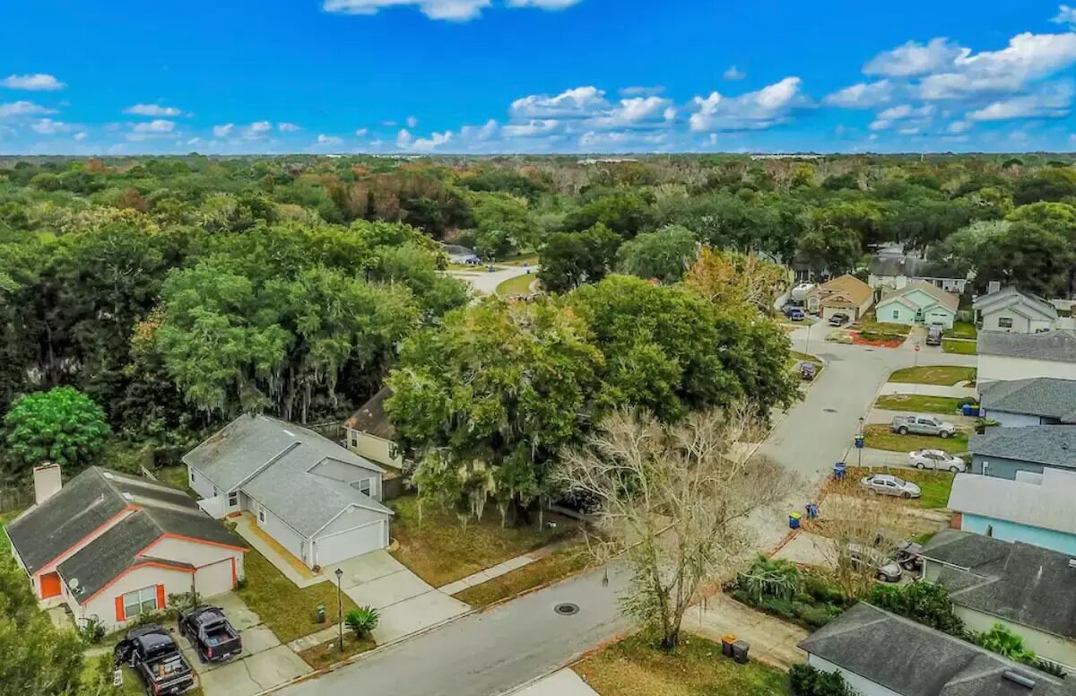 An aerial view of a residential area with houses, a central road, and surrounding greenery.