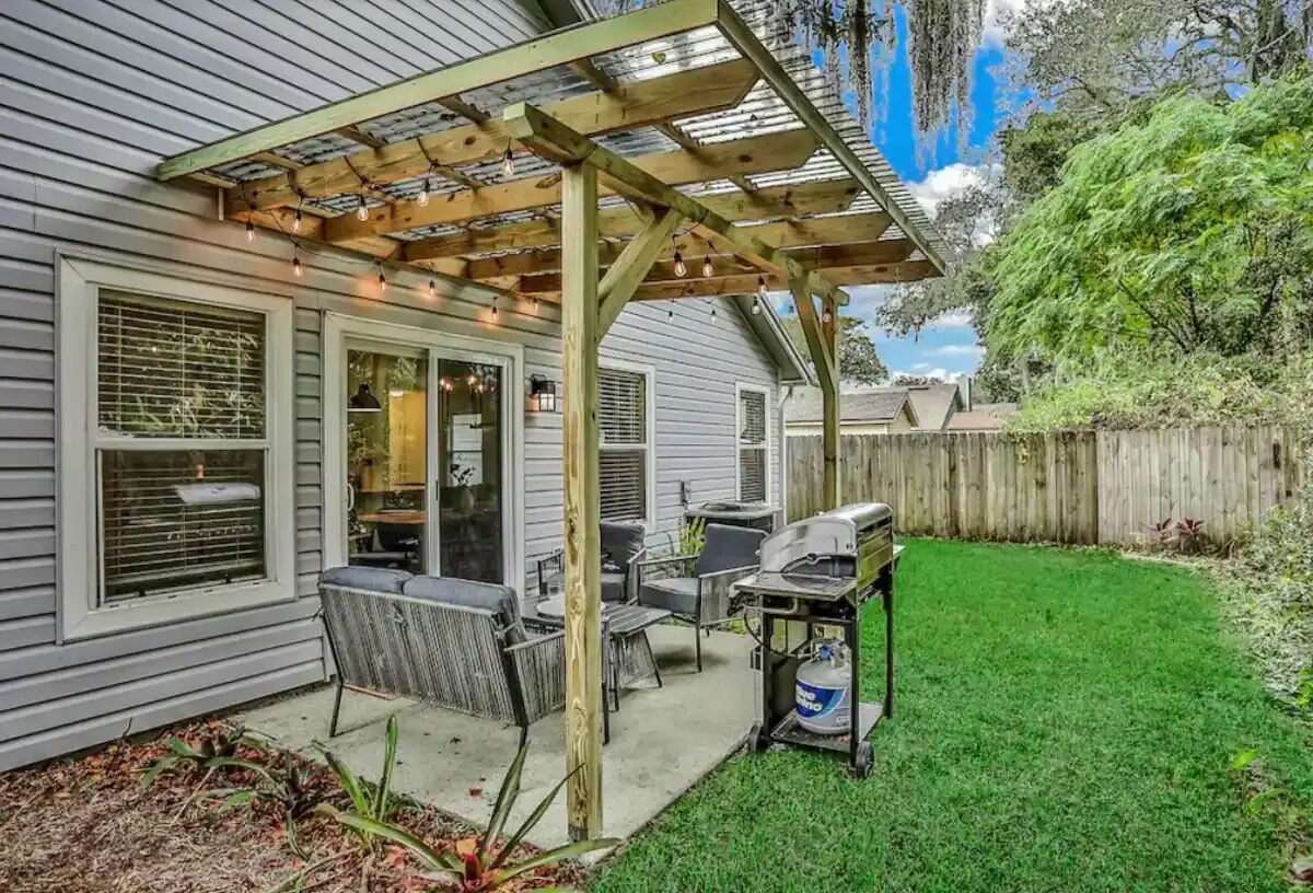 A covered patio with outdoor furniture, a grill, and a wooden fence in the background.