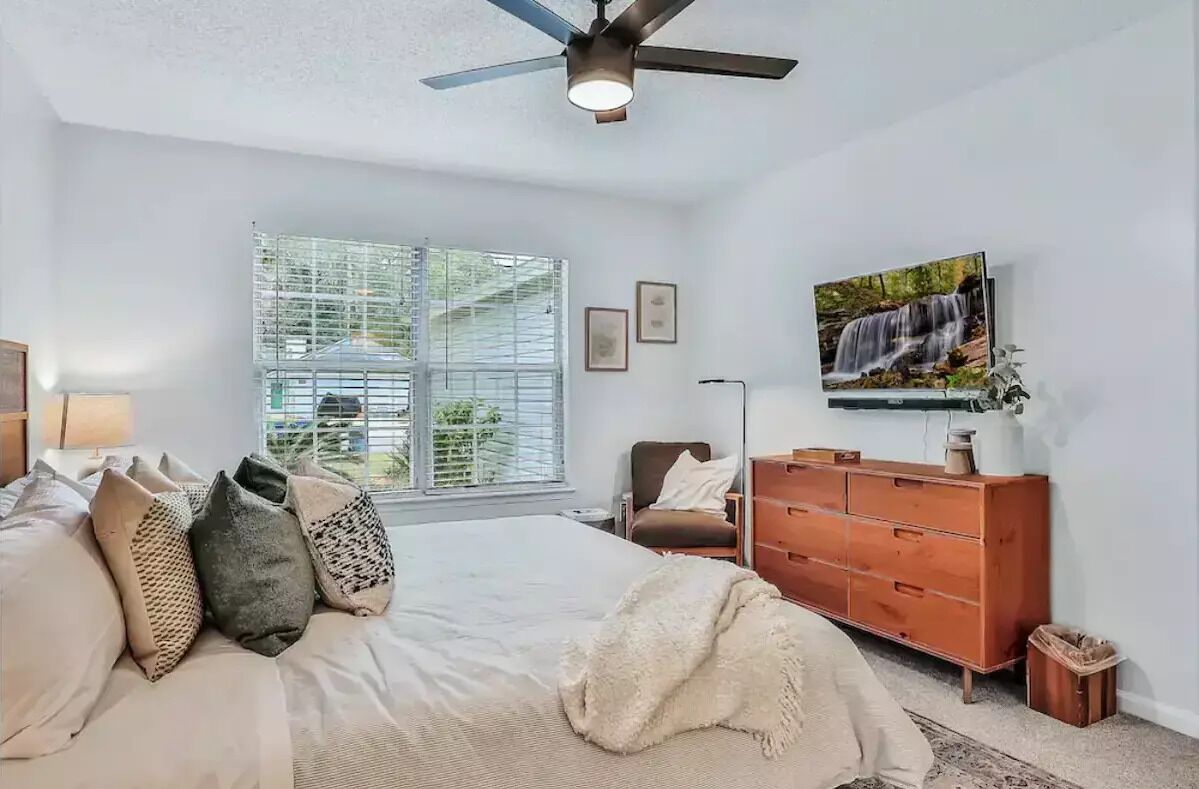 A bedroom with a bed, a dresser, a chair, a ceiling fan, a window with blinds, and a TV on the wall.